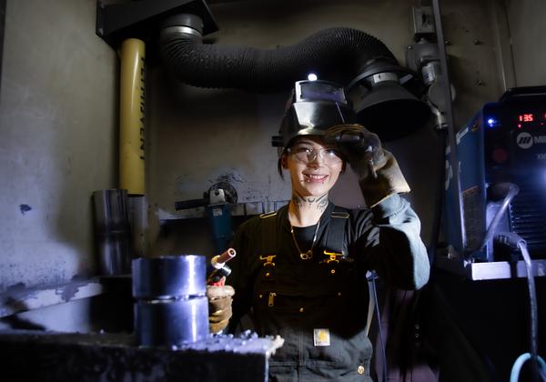 A female welding student has pushed her welding helmet up and is smiling at the camera. She is wearing black work dungarees, and safety glasses and is surrounded by welding equipment. 