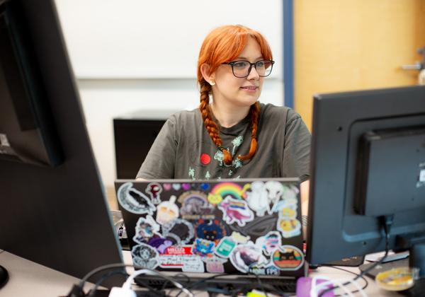 A male and a female web and mobile development students sit side by side working on their laptops. Between them is a potted plant related to a project they are working on together. 
