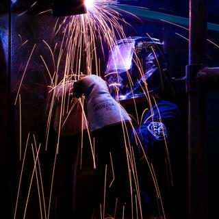 VIU student practices their welding techniques. Sparks are flying and their helmet is reflecting purple light.